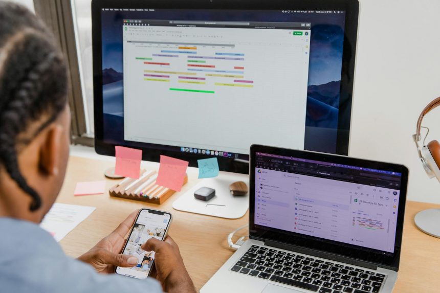 Businessman multitasks with smartphone, laptop, and monitor in office setting.