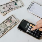From above of crop anonymous economist calculating total amount of income using calculator app on cellphone near piles of greenbacks and notebook with pen