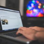 Hands typing on a laptop with a blog post visible, cozy indoor setting with colorful screen in background.